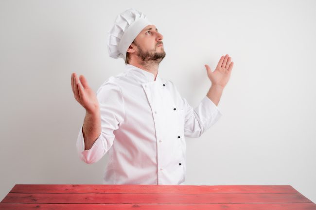 Young male chef in white uniform with open arms looking up, side confusing cooking instructions