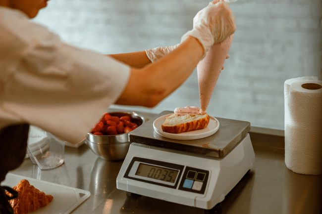 Confectioner applies pink cream onto croissant on scales in craft bakery shop food scales