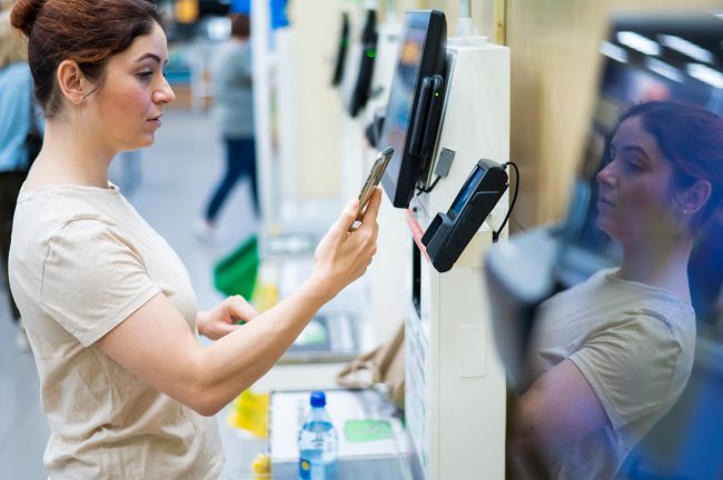 Woman paying with smartphone in store at self-checkout counter. self‑checkout overcharge