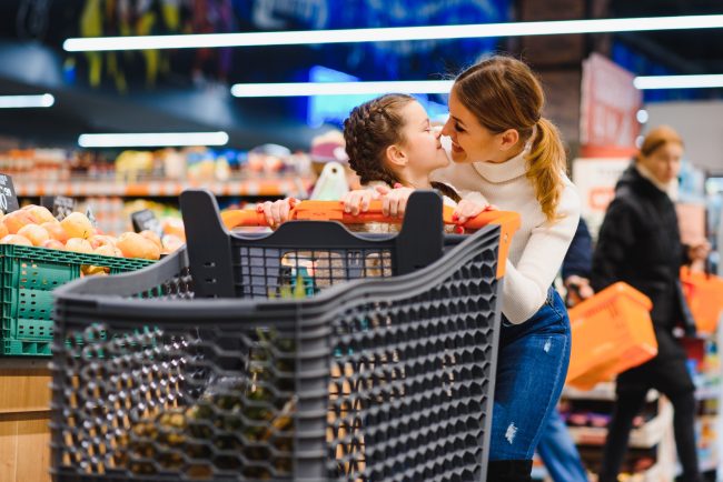 beautiful mother holding grocery basket with her child walking in supermarket. Shopping for healthy. cart tactics stores use to encourage overspending