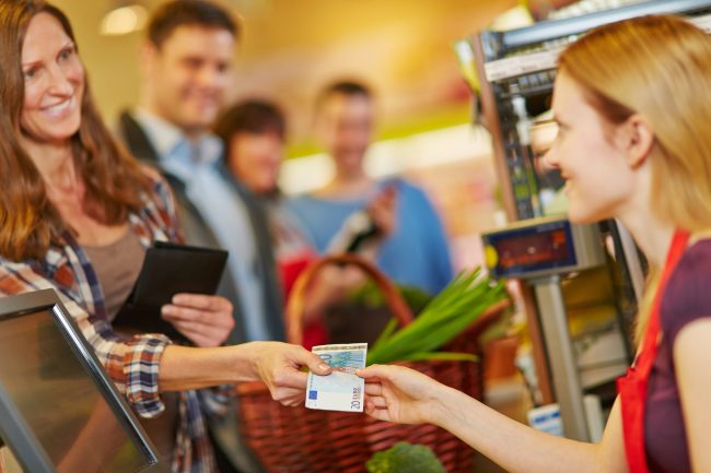 Woman paying with money at supermarket checkout Checkout Games: 8 Tricks Cashiers Use That Can Change Your Total Without You Noticing