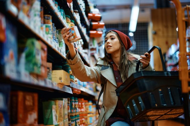 Young,Woman,Buying,Canned,Food,At,Grocery,Store. The Grocery Aisle That's Re-Arranged Monthly (And Why It's Designed to Confuse You)