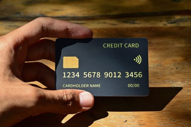 A hand holding a generic black credit card with a chip and contactless payment symbol over a wooden table. The Cash-Back Credit Card Categories Made for Grocery Shoppers