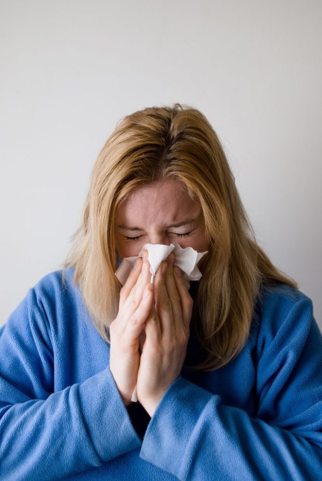 A woman in a blue sweater blowing her nose into a tissue. The “Allergy Shelf Check” Every Household Should Do Once a Month