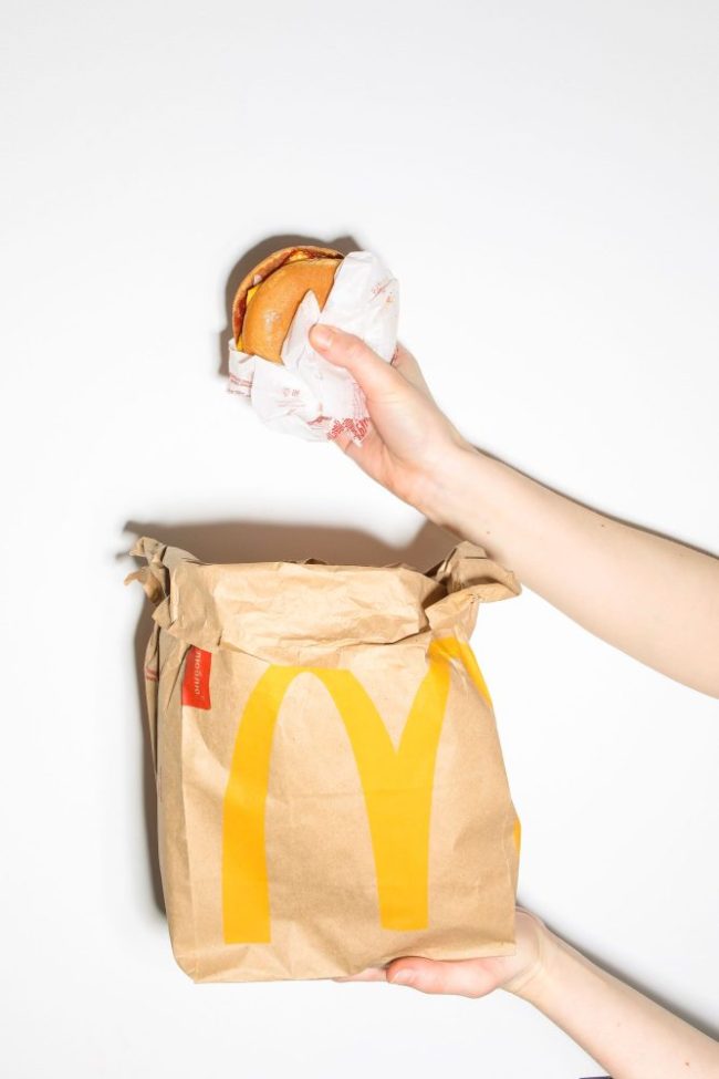 A hand lifting a wrapped burger out of a classic brown McDonald’s takeout bag. Why McDonald's Employees Assemble Every Burger the Same Way