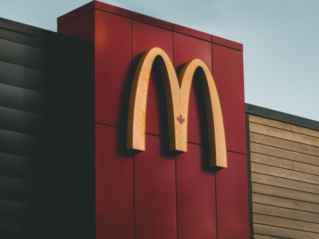 The glowing yellow golden arches logo mounted on a red exterior wall of a McDonald’s restaurant. This McDonald’s Secret Menu Hack Requires You to Buy 3 Sandwiches — Is It Worth It?