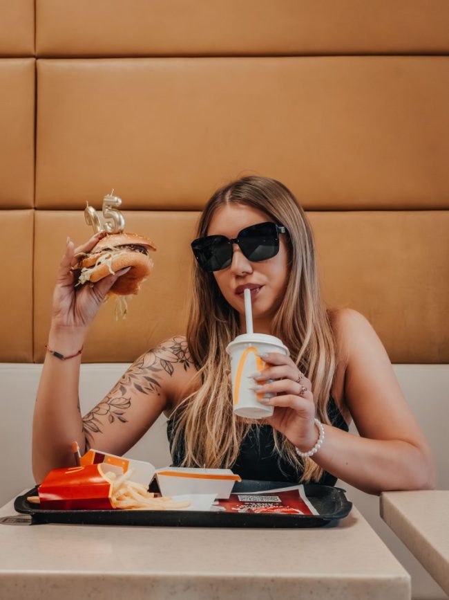 A woman wearing sunglasses holding a burger with a number 15 candle and drinking from a cup with a straw. The Same Big Mac Costs More Depending on Where You Live — We Found the Biggest Price Gaps