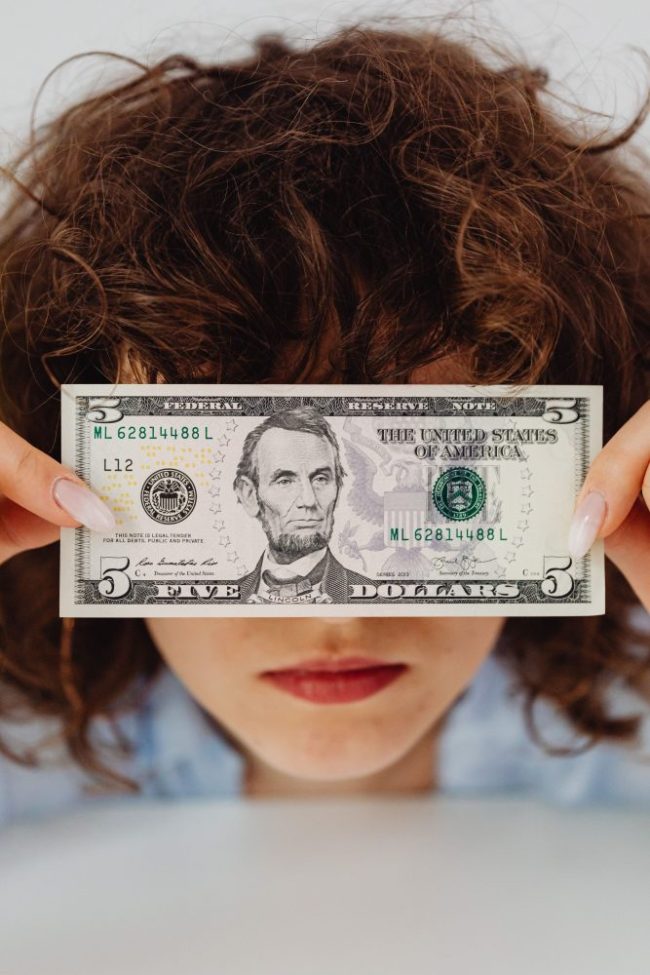 A person holding a five-dollar bill in front of their eyes. That Digital Coupon Says “Save $5 ” — Here’s Why It May Not Work at Checkout