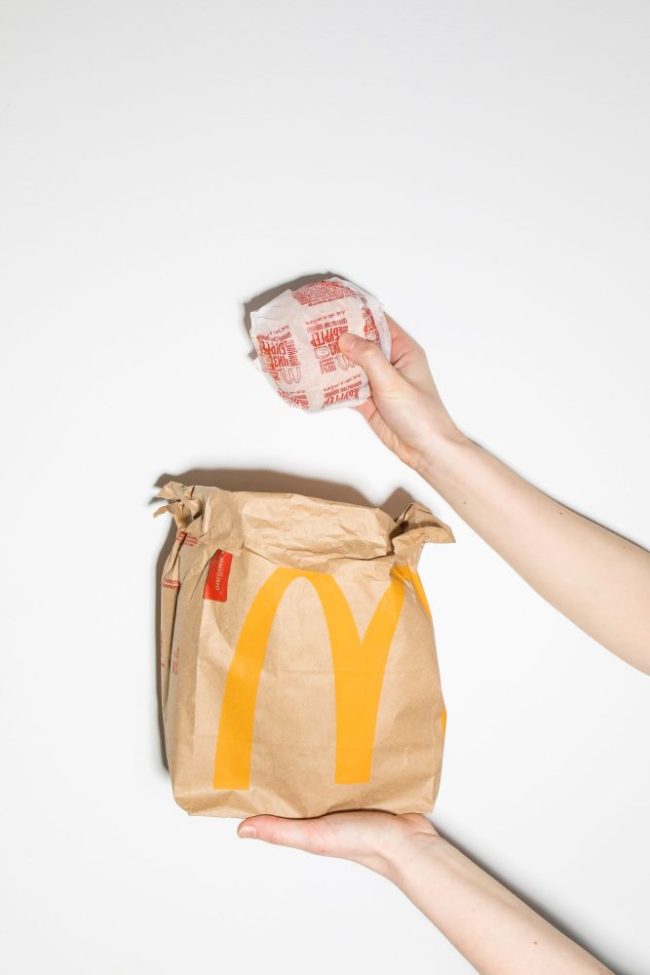 A person holding a wrapped burger above a McDonald’s takeout paper bag against a white background. McDonald’s, Wendy’s or Burger King: Which Value Meal Offers the Most Food for Your Money?
