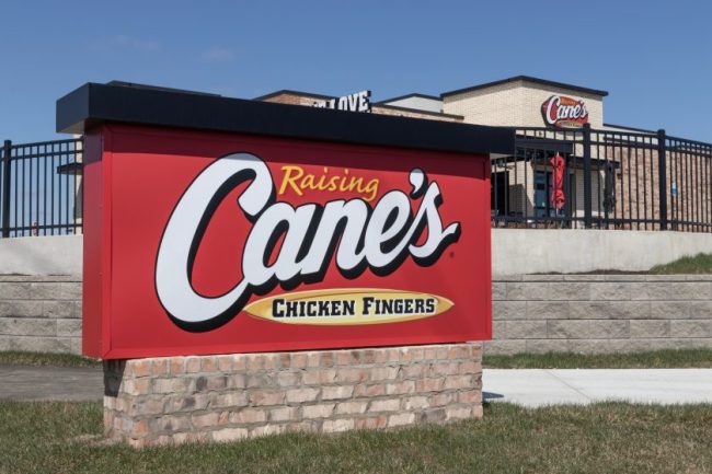 A red roadside sign for Raising Cane’s Chicken Fingers with the restaurant building behind it. The $15 Fast-Food Lunch Is Becoming Normal — Here’s What the Same Money Buys at 5 Chains