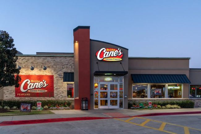 The exterior facade and lit sign of a Raising Cane’s Chicken Fingers restaurant. 8 Unwritten Rules to Know Before Ordering at Raising Cane’s