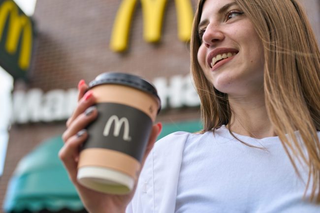 A smiling woman holding a McDonald’s coffee cup with the golden arches sign in the background. What Does $20 Buy at Costco’s Food Court vs. McDonald’s?