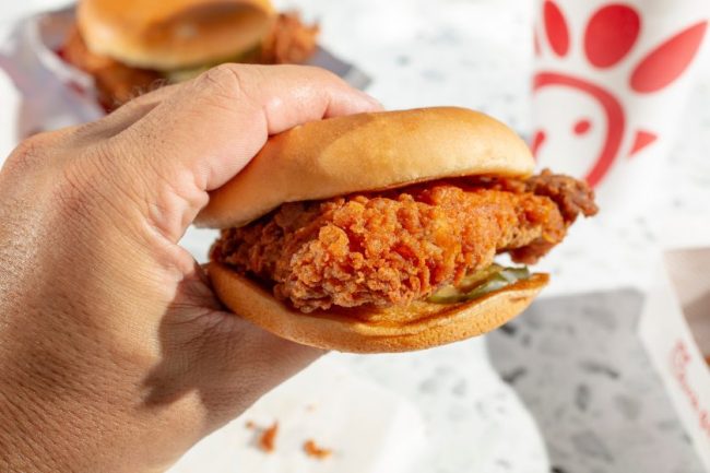 A person holding a crispy fried chicken sandwich in front of a fast-food branded cup. Is the Combo Meal Actually Cheaper? We Did the Math at 6 Fast-Food Chains