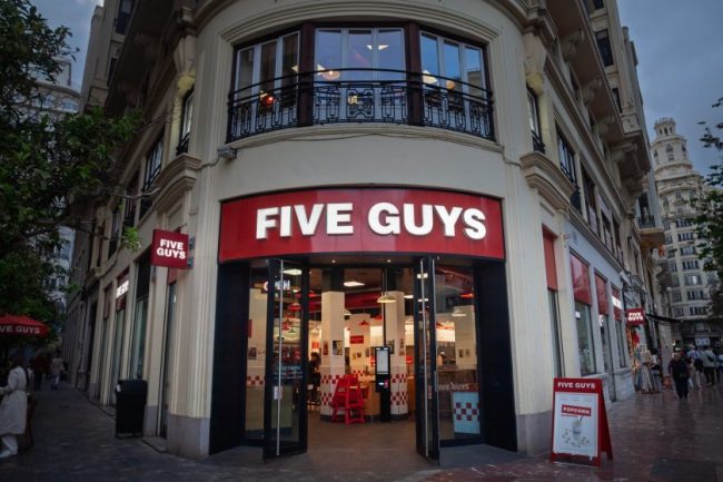 The exterior entrance and red signage of a multi-story Five Guys restaurant. 7 Fast-Food Sides That Cost Almost as Much as an Entire Value Meal